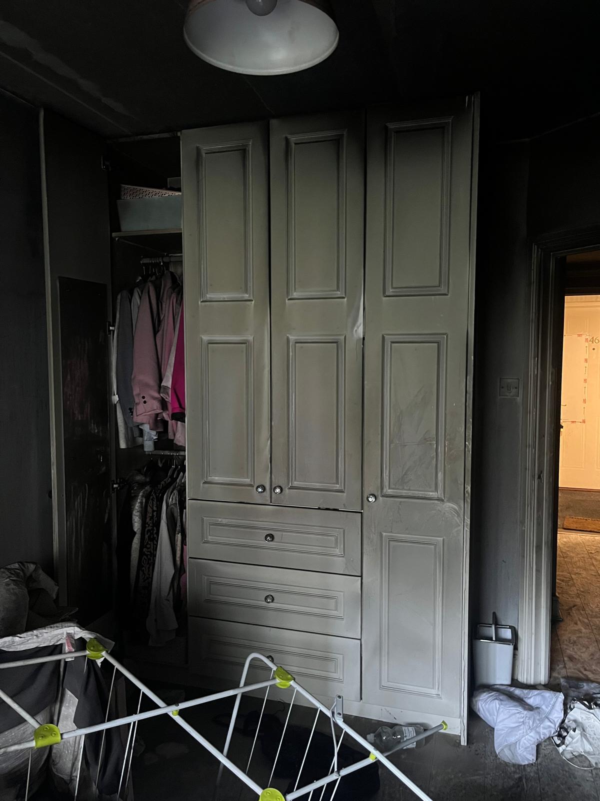 Fitted wardrobe scorched by fire, with smoke-blackened walls and a laundry rack on the floor